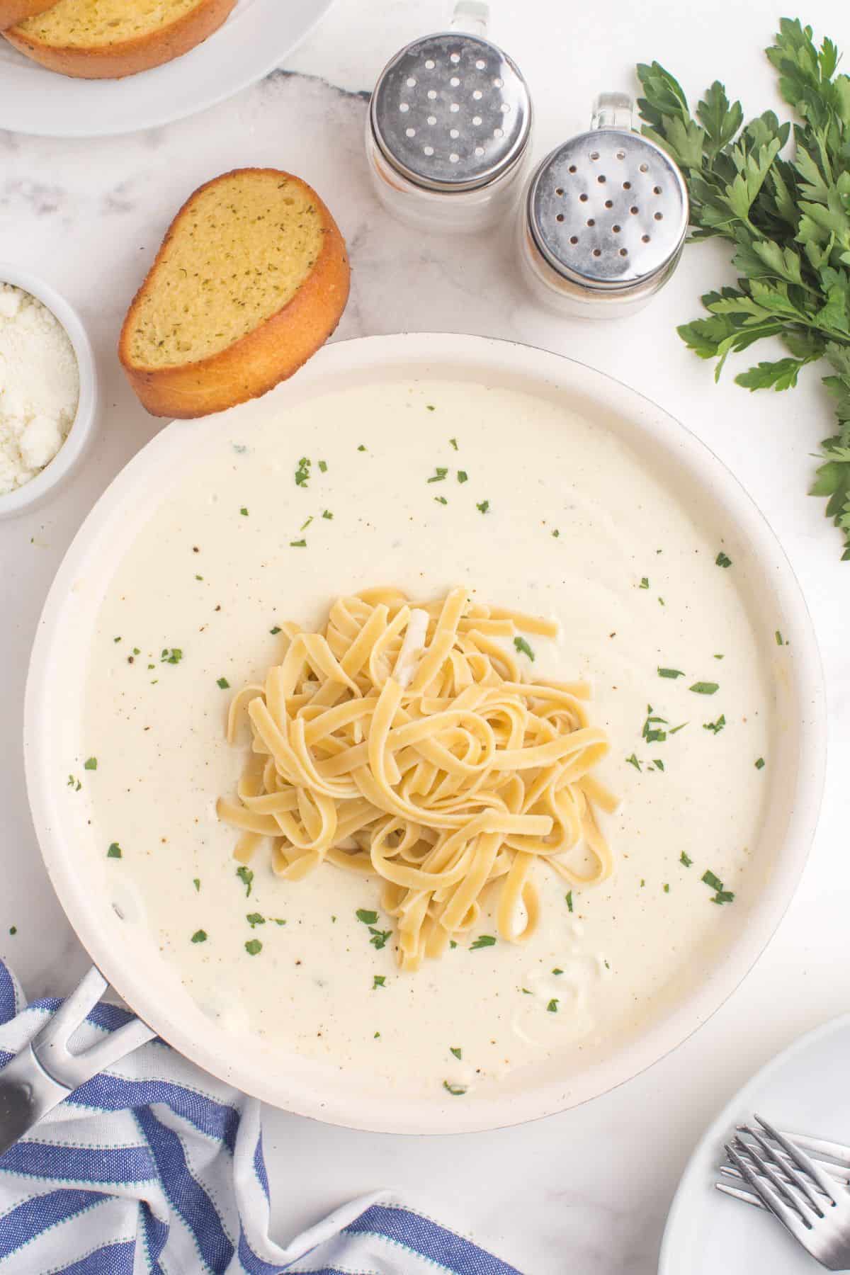 A pasta bowl with alfredo sauce. A pile of fettuccine pasta is piled in the center of the bowl. The pasta bowl is flanked by a slice of garlic bread, bowl of parmesan cheese, a plate and forks, parsley, and a set of salt and pepper shakers.