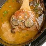 A wooden spoon lifting a cooked pork chop out of the crock pot.