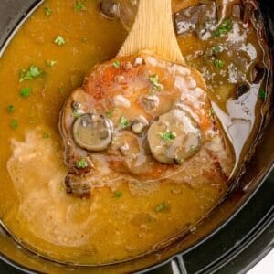 A wooden spoon lifting a cooked pork chop out of the crock pot.