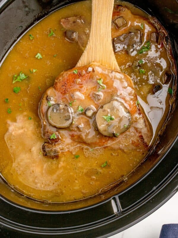 A wooden spoon lifting a cooked pork chop out of the crock pot.