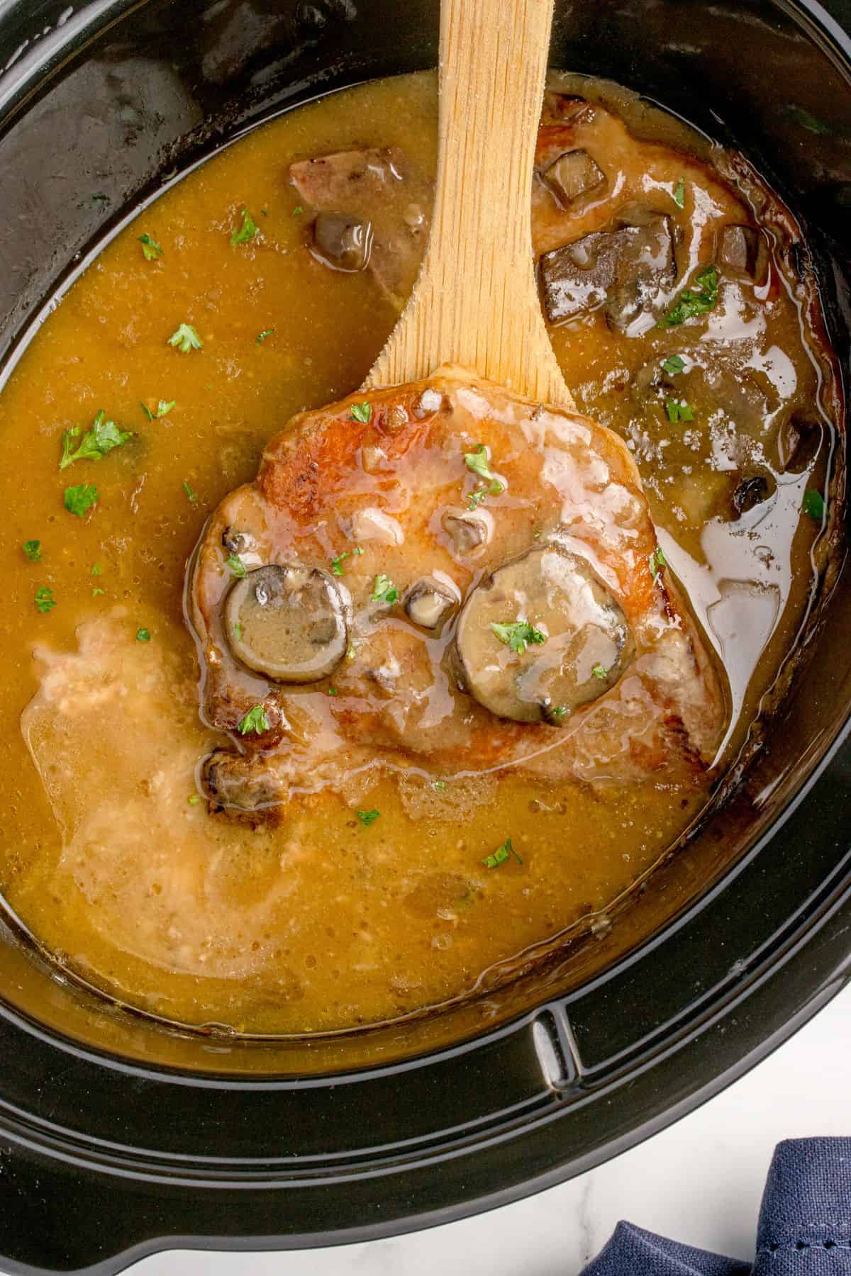 A wooden spoon lifting a cooked pork chop out of the crock pot.