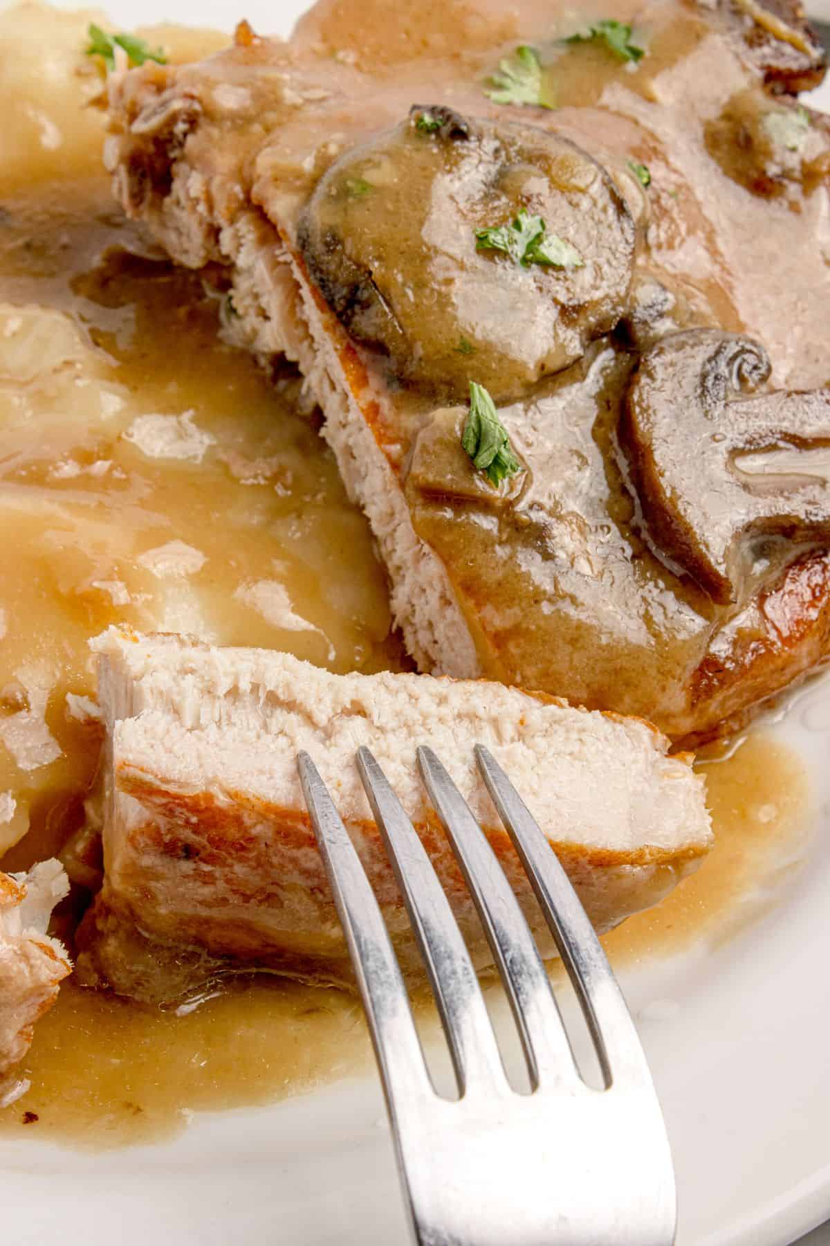 Sliced pork chop in mushroom gravy on a dinner plate. A fork is lifting the sliced pork chop to show the interior.