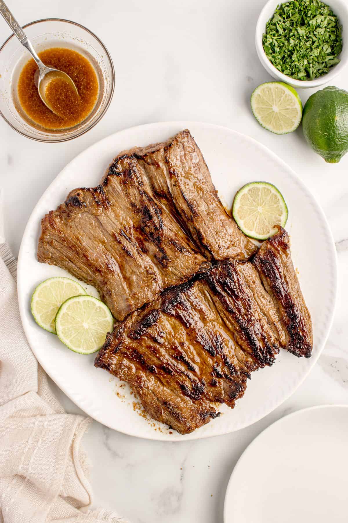 Completed skirt steak on a white dinner plate, garnished with lime slices. A small amount of marinade is in a glass jar to baste.