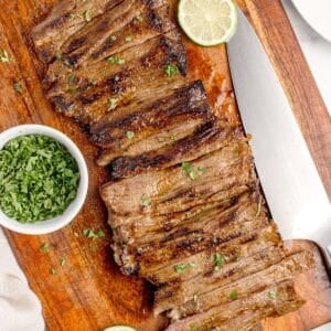 Sliced skirt steak on a wooden cutting board. Steak is surrounded by sliced limes and a small bowl of chopped parsley.