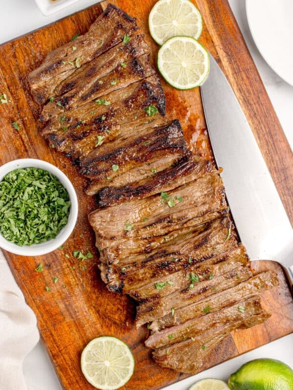 Sliced skirt steak on a wooden cutting board. Steak is surrounded by sliced limes and a small bowl of chopped parsley.