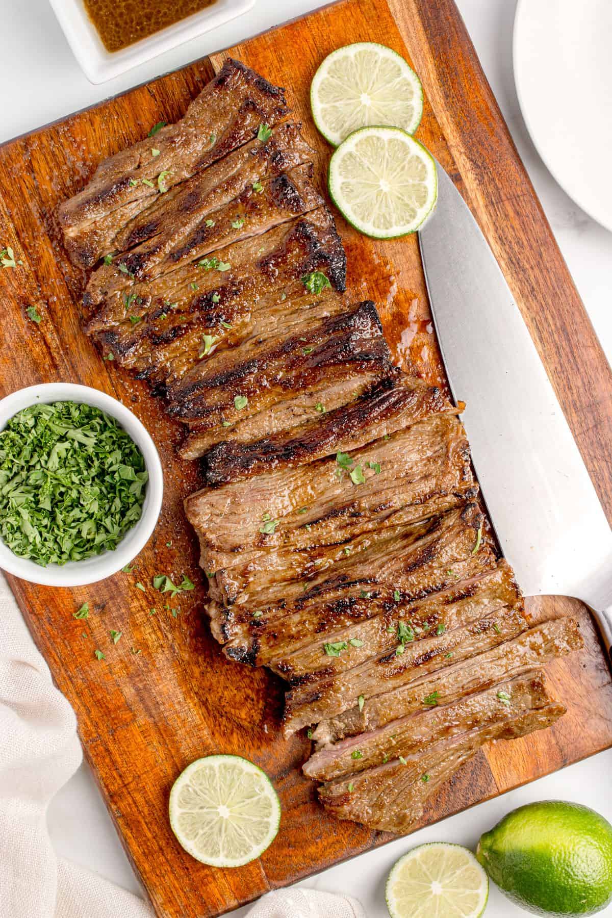 Sliced skirt steak on a wooden cutting board. Steak is surrounded by sliced limes and a small bowl of chopped parsley.