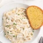 A white dinner plate with fettuccine alfredo and a slice of garlic bread. A large skillet of alfredo sauce is next to the plate.