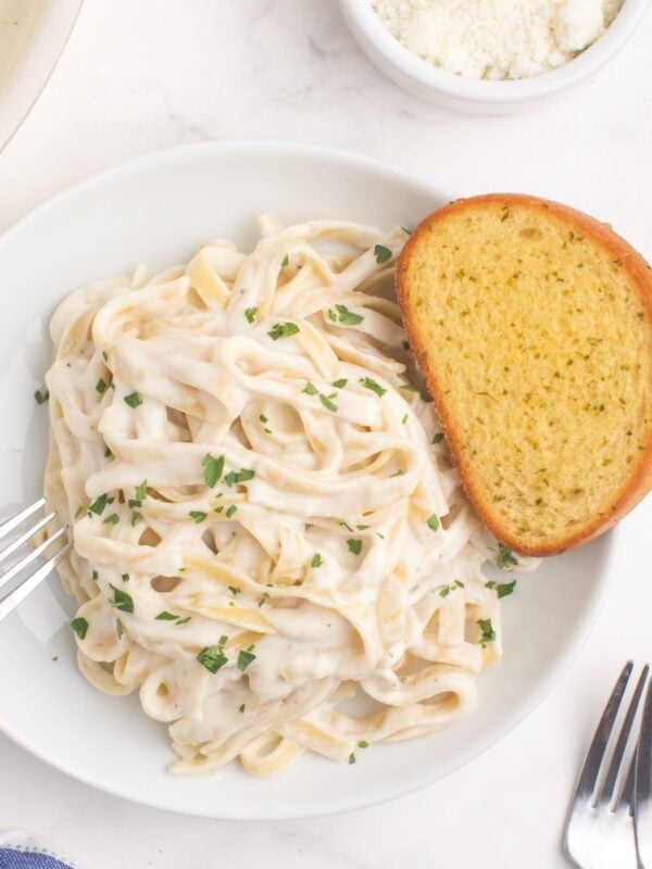 A white dinner plate with fettuccine alfredo and a slice of garlic bread. A large skillet of alfredo sauce is next to the plate.