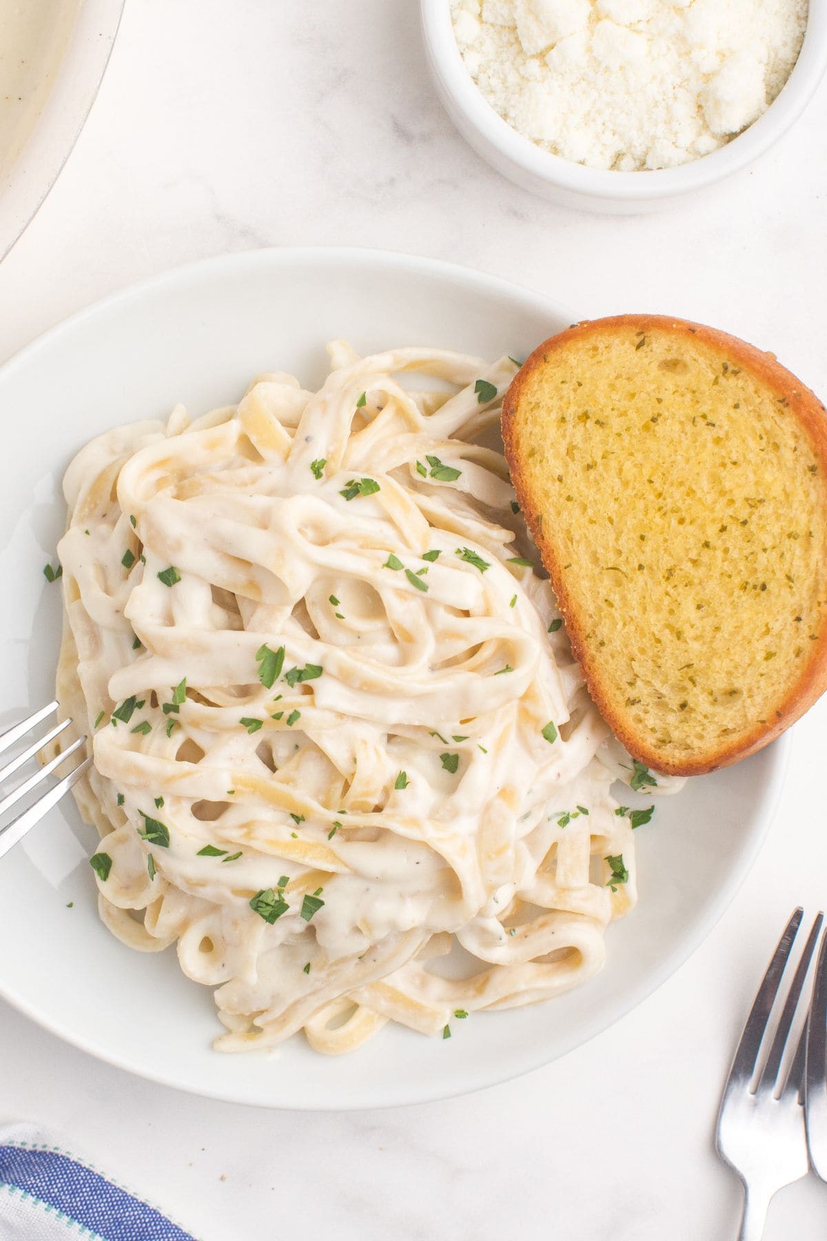 A white dinner plate with fettuccine alfredo and a slice of garlic bread. A large skillet of alfredo sauce is next to the plate.