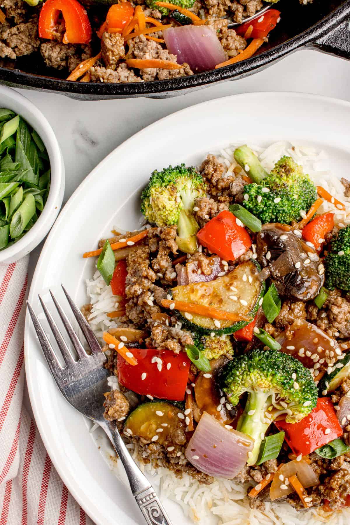 A serving of stir fry on a white dinner plate. A skillet with the remaining stir fry is in a cast iron pan above the plate.