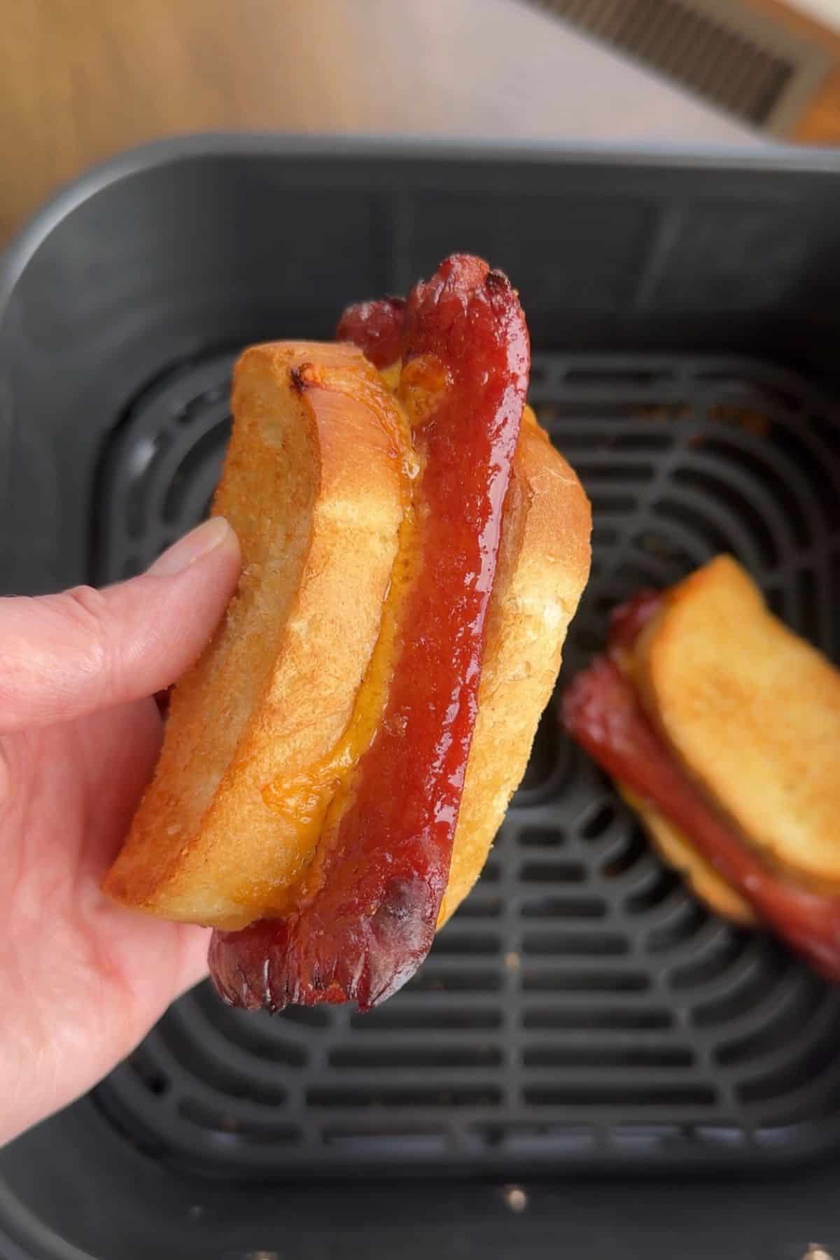 Air fryer grilled cheese hot dog being held above the air fryer basket to see the side all cooked and delicious.