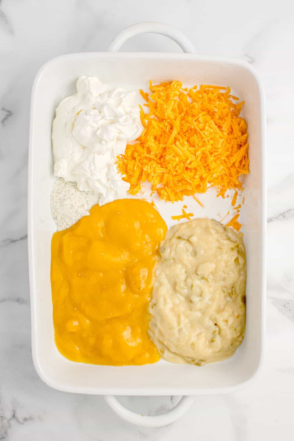 White baking dish with cream of chicken soup, cream of celery soup, sour cream, milk, ranch seasoning, and cheddar cheese combined.